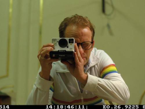 Ein Mann in einer weißen Jacke mit rot-orange-gelb-grün-blauen Streifen an Armen und über der Brust steht vorübergebeugt. In den Händen eine Polaroid-Kamera, er guckt durch den Sucher. Am unteren Bildrand die Bildinformation eines Polaroidbildes.
